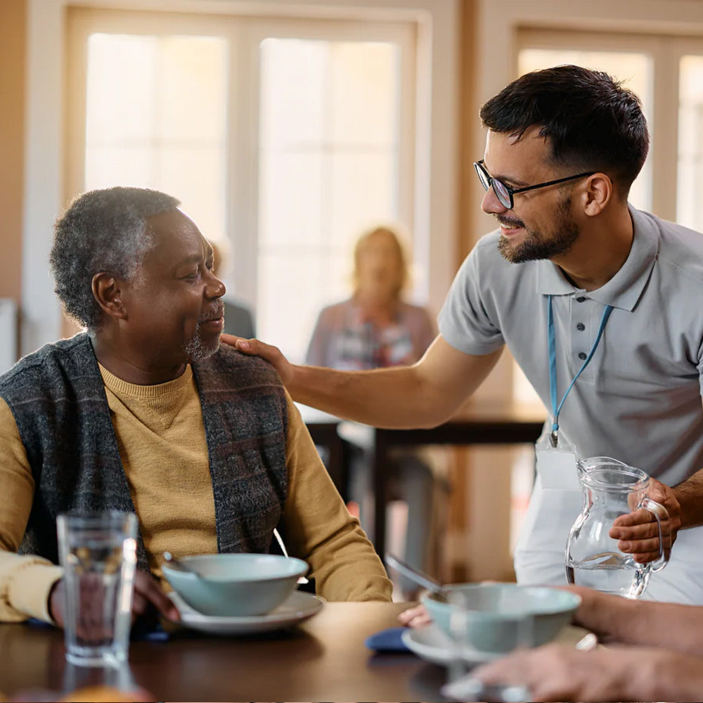 Care worker supporting elderly man at table, supplements health, bright indoor setting